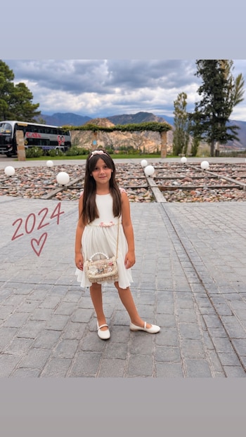 Una niña con vestido blanco y bolso pequeño posa de pie en un piso empedrado con un paisaje de montañas al fondo