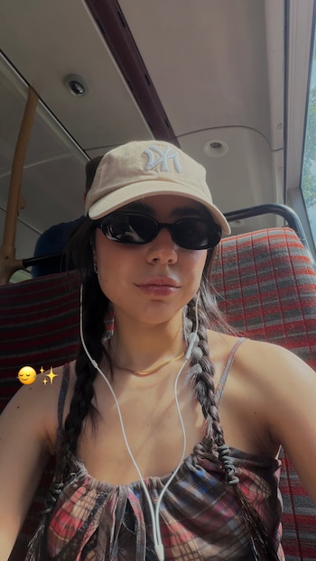 Una mujer joven con gorra beige, lentes de sol oscuros, trenzas y auriculares blancos está sentada en un autobús. El asiento es de tela a cuadros rojos y grises