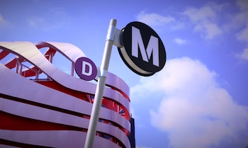 Señal vertical con dos carteles redondos: uno negro con una 'M' blanca y otro morado con una 'D' blanca, junto a un edificio moderno con fachada ondulada