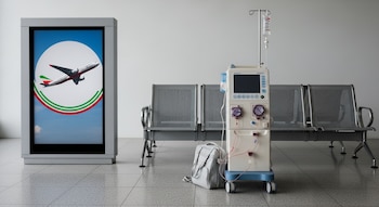 Máquina de diálisis portátil, maleta médica y sillas de espera grises en un aeropuerto vacío. Un cartel luminoso muestra un avión blanco con franjas verde y rojo.