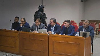 Seis hombres con traje sentados frente a computadoras en una mesa de madera y un agente de seguridad armado detrás de ellos