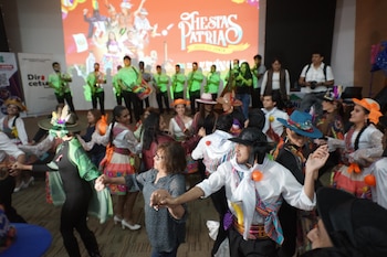 Grupo de personas bailando con vestimenta festiva y sombreros. Pantalla de fondo con el texto Fiestas Patrias. Varias personas observan el evento