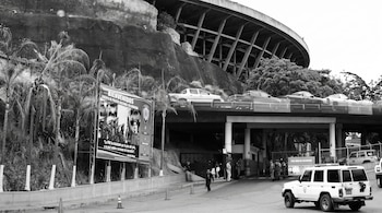 Fotografía en blanco y negro de una estructura de concreto con un cartel de la Policía Nacional Bolivariana, patrullas y guardias.