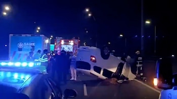 Coche blanco volcado sobre la calzada, ambulancia con logo SAME, camión de bomberos, luces de emergencia azules, varios operarios y farolas en la oscuridad