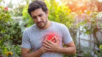 Hombre con camiseta gris sostiene su pecho con ambas manos; el área del corazón está resaltada en rojo; fondo verde borroso con vegetación.
