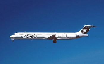 Avión comercial blanco con el nombre Alaska en el fuselaje y la cara de un hombre en la cola, sobre un fondo de cielo azul