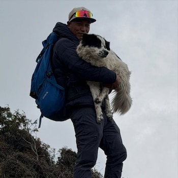 Hombre con chaqueta oscura, gorra, lentes de sol y mochila azul carga un perro grande, blanco y negro. Detrás, cielo nublado y vegetación