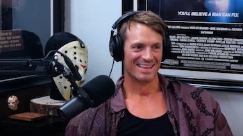 Joel Kinnaman, con auriculares y camisa estampada, sonríe frente a un micrófono. Detrás, una máscara de hockey y un póster de película son visibles