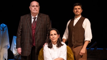 Una mujer sentada vestida de blanco está flanqueada por dos hombres con trajes elegantes en un escenario teatral de fondo oscuro