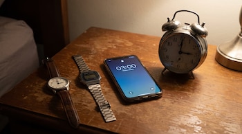 Un reloj analógico con correa de cuero, uno digital de metal, un teléfono móvil y un despertador sobre una mesa de madera marcan las 03:00.