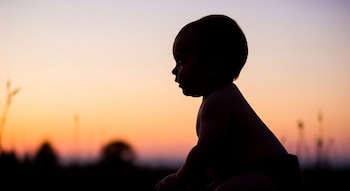 Silueta de un bebé de ocho meses sentado de perfil, mirando hacia la izquierda, contra un cielo anaranjado, rosa y morado durante el atardecer, con flora difusa en el fondo.