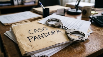 Un expediente de cartón con el texto CASO PANDORA, hojas de papel y unas esposas metálicas sobre un escritorio de madera.