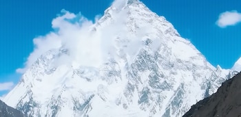 Una montaña cubierta de nieve y hielo se eleva bajo un cielo azul con nubes blancas. Rocas oscuras en la parte inferior derecha