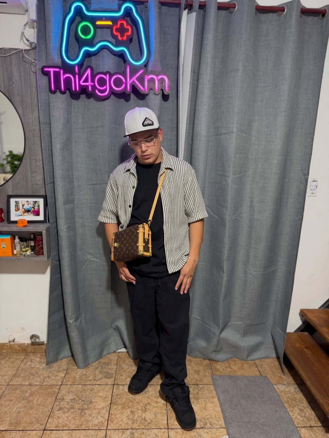 Thiago Medina posa con un atuendo casual que incluye una camisa a rayas, gorra blanca y un bolso cruzado de Louis Vuitton, destacando su estilo personal frente a un letrero de neón.