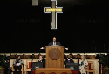 Hombre en traje habla desde un púlpito de madera. Detrás, una cruz de neón con 'SUNSHINE BAND' y un fondo negro con las palabras 'I AM A MAN'