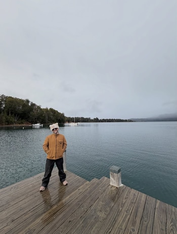 Un hombre con chaqueta marrón, gorro de piel y gafas de sol posa de pie en un muelle de madera con el lago y árboles al fondo