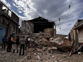 Hombres de pie sobre una calle de adoquines entre edificios en ruinas y montones de escombros bajo un cielo nublado con cables eléctricos