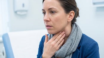Mujer de cabello oscuro con chaqueta azul y bufanda gris se toca el lado derecho del cuello con la mano izquierda en un entorno médico.