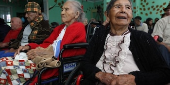 Dos mujeres mayores en sillas de ruedas se encuentran sentadas en primer plano junto a otros adultos al fondo de la habitación
