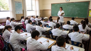 Una maestra con guardapolvo blanco da clase a estudiantes con guardapolvos blancos en un aula con pupitres, pizarrón, ventanas y carteles.