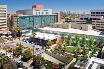 Vista aérea del Children's Hospital Los Angeles: edificio moderno de varios pisos con fachada de vidrio y beige, rodeado de calles, palmeras y vegetación. Fondo urbano