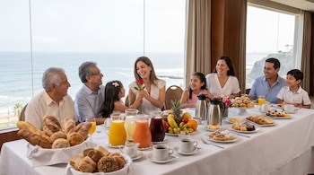 Una familia de varias generaciones disfruta un desayuno buffet con vista al mar. La mesa tiene panes, frutas, jugos y dulces. Una madre recibe una rosa.