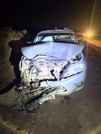 Camioneta gris con daños graves en la parte frontal, la carrocería y el capó abollados, en un camino de tierra durante la noche, con luces al fondo