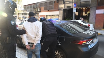 Dos agentes de policía, uno sujetando a un hombre esposado y otro junto a un vehículo policial negro con luces azules, en una calle urbana