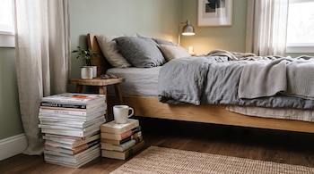 Pila de revistas, libros con una taza blanca encima, cama de madera con sábanas grises, alfombra de fibra y mesa auxiliar con planta.