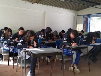 Estudiantes con chaquetas oscuras escriben en sus pupitres dentro de un aula con paredes de ladrillo blanco