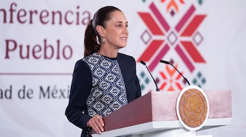 Mujer con saco oscuro y cabello recogido habla en un atril con escudo mexicano y dos micrófonos. Fondo blanco con texto parcial y patrón geométrico.