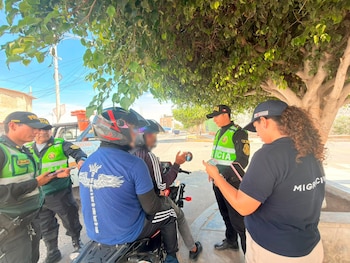 Agentes policiales y una funcionaria de Migraciones solicitan documentos a dos personas en una motocicleta bajo la sombra de un árbol