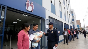Pareja con bebé en brazos y un agente uniformado frente a un edificio con logo de dos figuras y una huella dactilar. Fila de personas afuera.