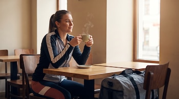 Una mujer con ropa deportiva sostiene una taza humeante en una mesa de madera junto a una ventana y una bolsa de gimnasio.