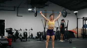 Mujer musculosa, Tia-Clair Toomey, levanta una barra con discos negros sobre su cabeza en un gimnasio. Viste top deportivo amarillo y shorts morados. Al fondo, equipo de gym