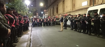 Vista de calle de noche con dos filas de hombres y mujeres uniformados, chalecos reflectantes naranjas, gorras, un hombre al centro, vehículos y edificios