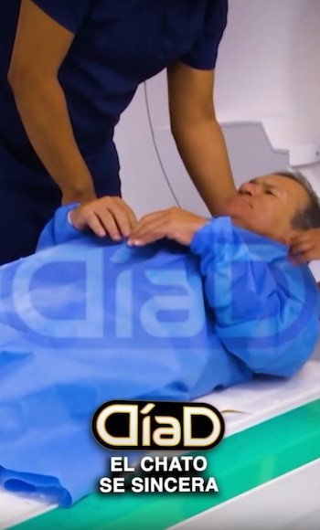 Un hombre con bata azul permanece recostado en la camilla de un escáner médico mientras una persona con uniforme azul lo asiste