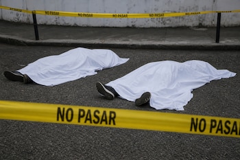Dos cuerpos cubiertos por sábanas blancas permanecen sobre el asfalto detrás de una cinta de seguridad amarilla con la leyenda NO PASAR.