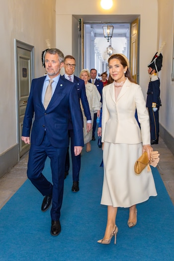 El Rey Federico X en traje azul marino y la Reina Mary con un traje crema y zapatos de tacón con estampado de serpiente, caminan por un pasillo