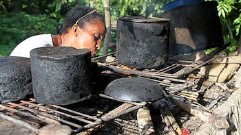Mujer con blusa blanca y pañuelo en la cabeza cocina en un fogón rústico al aire libre. Varias ollas negras descansan sobre una parrilla con fuego y humo