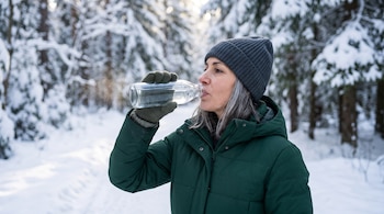 Una mujer con abrigo verde y gorro gris bebe agua de una botella de vidrio en un bosque lleno de nieve.