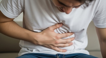Vista cercana del torso de una persona, vestida con camiseta blanca y jeans, inclinada con una mano sobre su abdomen.