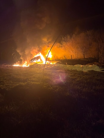 Imagen nocturna de un incendio con grandes llamas y humo, que consume una estructura. Se observa césped en primer plano y árboles en el fondo