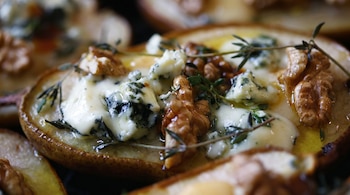 Primer plano de una pera asada cortada por la mitad, cubierta con trozos de queso azul cremoso, nueces enteras y rociada con miel, sobre un fondo desenfocado.