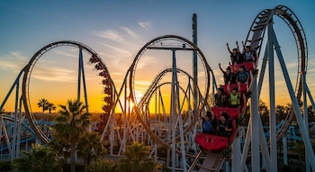 Una montaña rusa roja llena de personas con las manos en alto sube una curva, con el sol poniente y un cielo naranja y azul de fondo, entre palmeras.