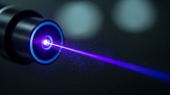 Primer plano de un dispositivo láser oscuro emitiendo un haz de luz violeta intenso con partículas dispersas a lo largo de su trayectoria. Un anillo azul rodea el emisor.