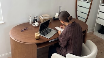 Mujer sentada de espaldas en un escritorio de madera con portátil, gafas, bebida, libros, vela y accesorios de oficina.