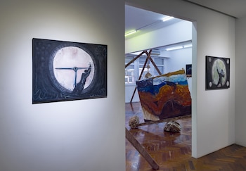Galería de arte con paredes blancas y suelo de parqué. Una pintura de reloj con figura humana en primer plano. Al fondo, otra pintura y una instalación de madera