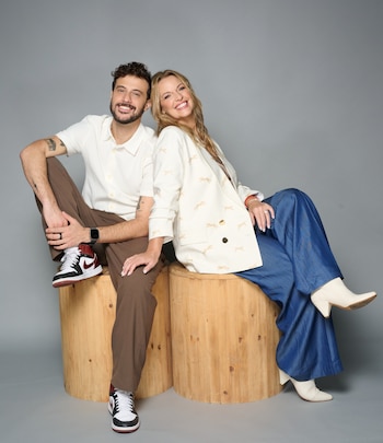 Diego Leuco y Carina Zampini sentados de espaldas en taburetes de madera, sonriendo a la cámara, con ropa informal de tonos claros y oscuros