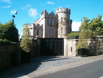 Un castillo de piedra gris con torreones y un gran portón negro en primer plano, flanqueado por muros de piedra y árboles verdes bajo un cielo azul